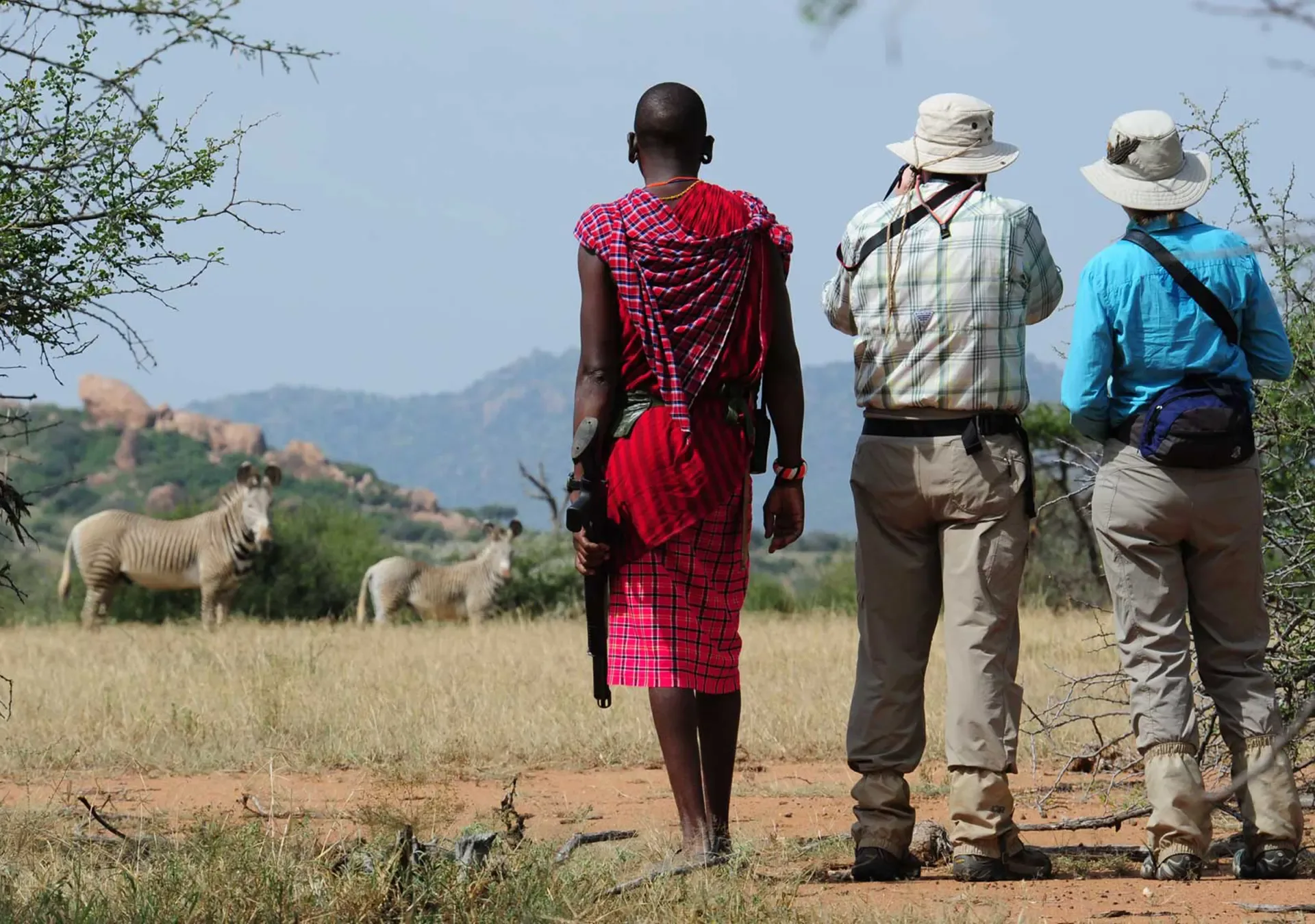 A Maasai guide and two guests watch zebras graze on the plains of Kenya.