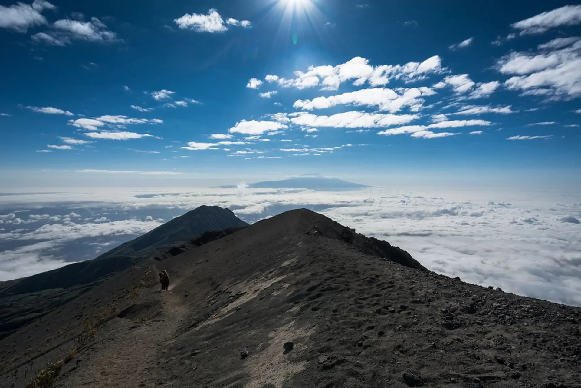 A hiker on the rocky ridge of Mt Meru, Tanzania, with the summit cone ahead and cloud filling the valley below.