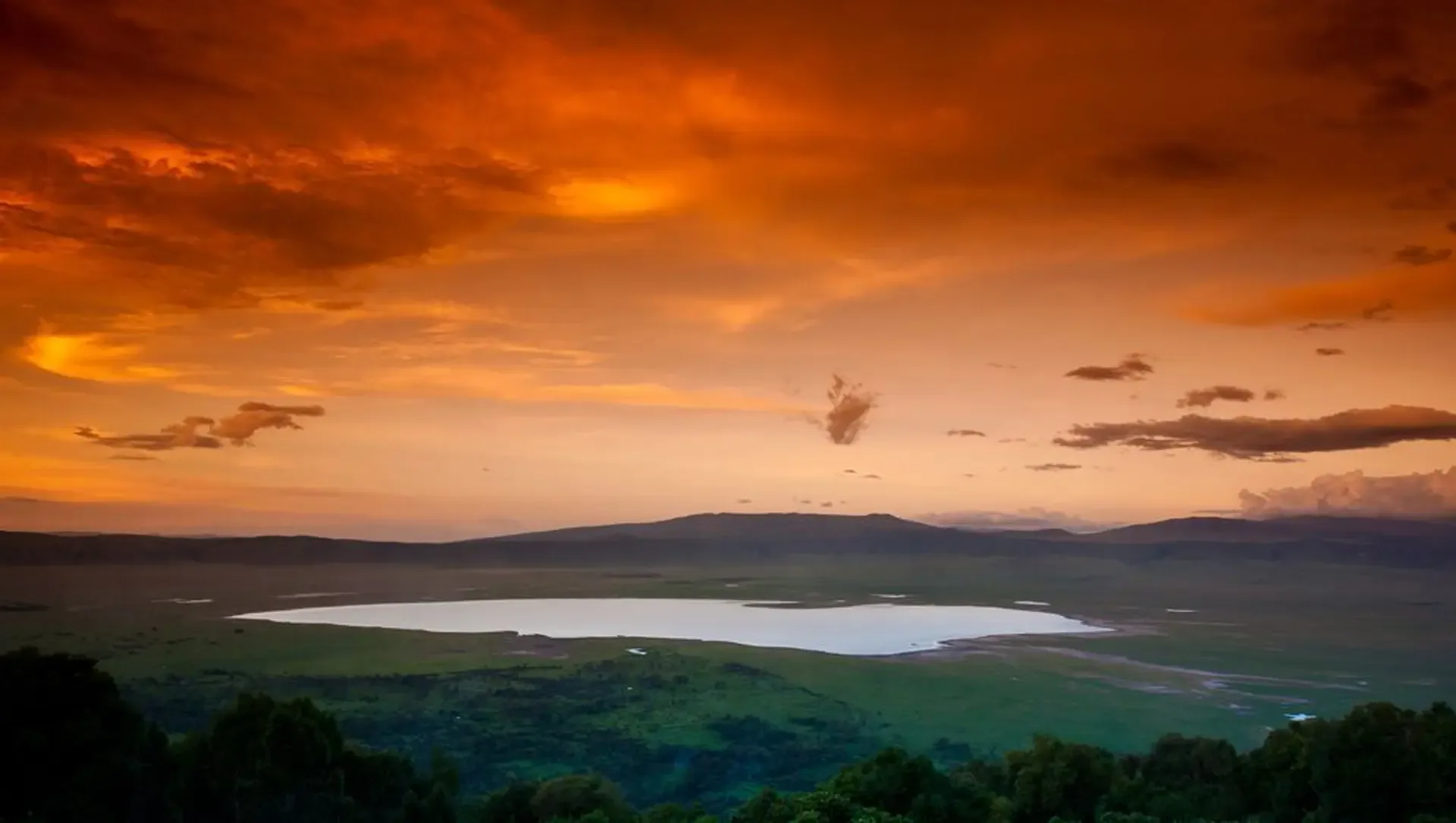 Dusk on the Ngorongoro Crater rim, with an orange sky over green highlands and a pale lake below.