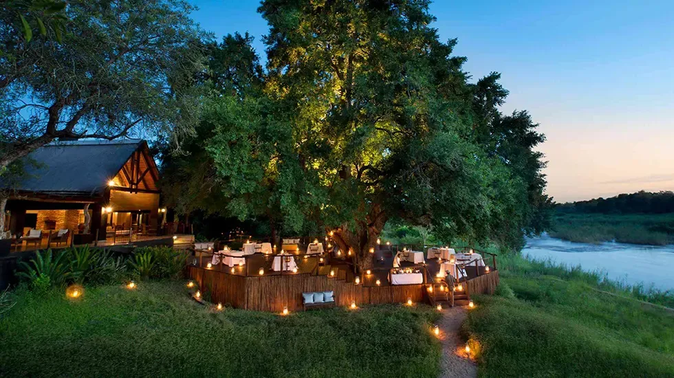 Dinner laid out beneath a lantern-lit tree at a Sabi Sands lodge, with the river beyond.