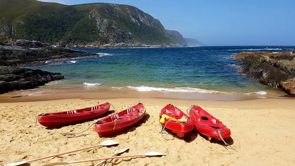 Red kayaks resting on the sand at Tsitsikamma, South Africa, beside calm turquoise water.