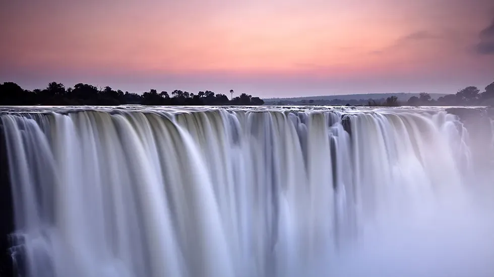 Sheer curtains of water at Victoria Falls, Zimbabwe, under a soft pink evening sky.