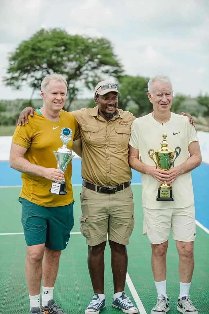 A Gosheni guide standing between two players holding trophies on a tennis court in the bush